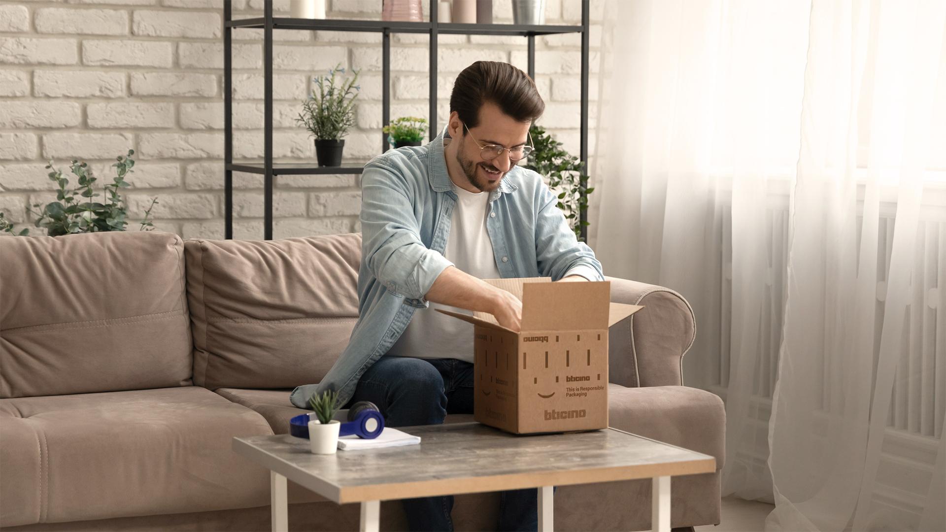 Un uomo seduto sul divano sorride mentre apre una scatola BTicino.